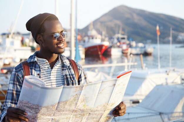 Concepto de personas, estilo de vida, viajes y turismo. Guapo joven turista afroamericano de moda vistiendo gafas de sol, sombrero y mochila estudiando el mapa de papel durante sus vacaciones en la ciudad europea