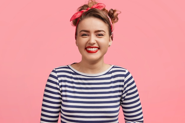 Concepto de personas y felicidad. Adorable joven sonriente vestida con un suéter de marinero, complacida por una historia agradable, se encuentra contra la pared rosa. Chica pinup alegre expresa positividad