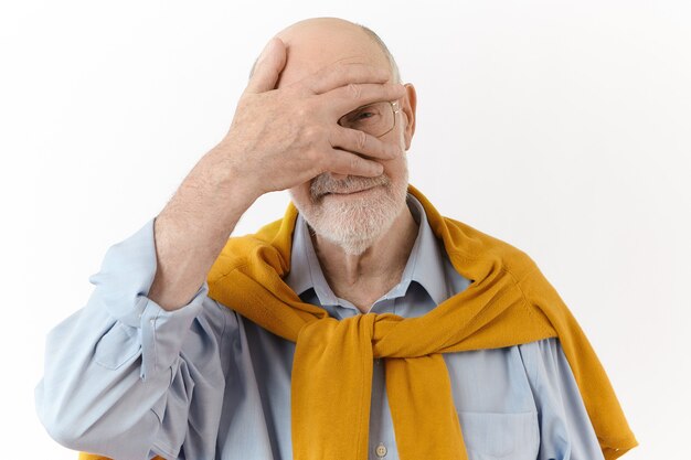 Concepto de personas, gestos y signos. Anciano caucásico elegante sin afeitar con anteojos y ropa elegante manteniendo la palma en la cara y mirando a la cámara a través de los dedos, posando aislado