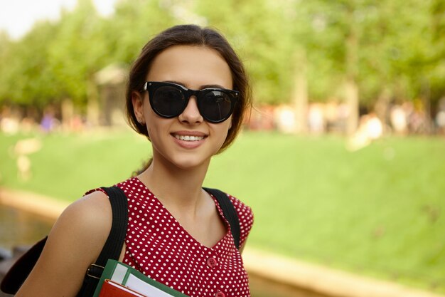 Concepto de personas, juventud, escuela y educación. Moda chica universitaria positiva feliz vistiendo tonos negros y llevando mochila disfrutando de un agradable clima de verano, yendo a casa de clases,