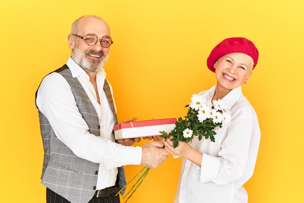 Concepto de personas maduras, edad, citas, romance y relaciones. Emocionada confundida mujer de mediana edad en el capó rojo sintiéndose incómoda mientras recibe un regalo inesperado y flores de un hombre barbudo mayor