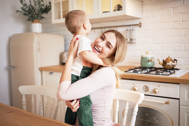 Concepto de personas, maternidad, amor, familia y relaciones. Retrato de mujer joven y bonita feliz sentada en el interior de la cocina elegante abrazando a su adorable hijo, mirando con sonrisa alegre