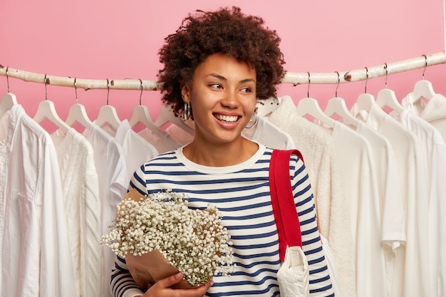 Concepto de personas, moda y consumismo. Feliz mujer étnica mira hacia otro lado, vestida con jersey de rayas, lleva ramo y bolso