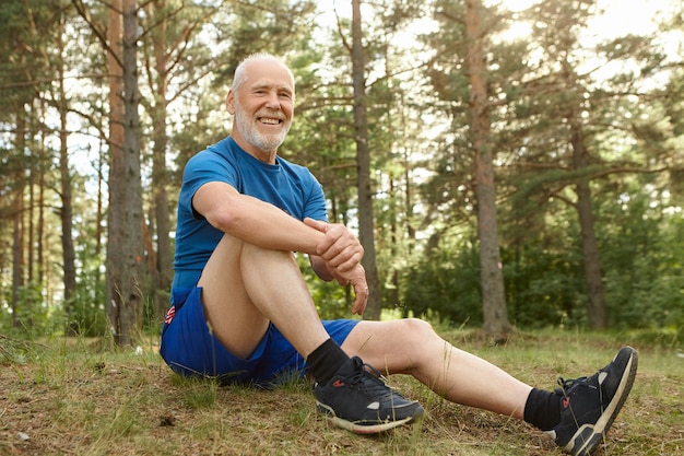 Concepto de personas, naturaleza, deportes y ocio. Feliz hombre jubilado sin preocupaciones con barba gris sentado cómodamente en la hierba en el bosque de pinos, manteniendo el codo en la rodilla, descansando después del ejercicio cardiovascular al aire libre