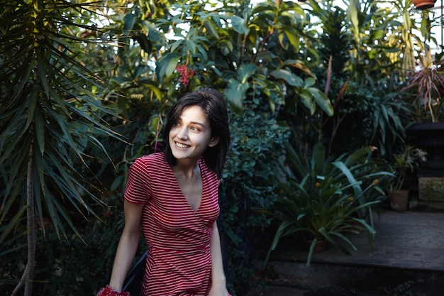 Concepto de personas, naturaleza y enivronment. alegre joven de cabello oscuro con una sonrisa encantadora relajándose al aire libre, respirando aire fresco entre las plantas tropicales, con expresión feliz en su cara bonita