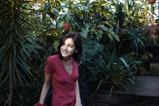 Concepto de personas, naturaleza y enivronment. Alegre joven de cabello oscuro con una sonrisa encantadora relajándose al aire libre, respirando aire fresco entre las plantas tropicales, con expresión feliz en su cara bonita