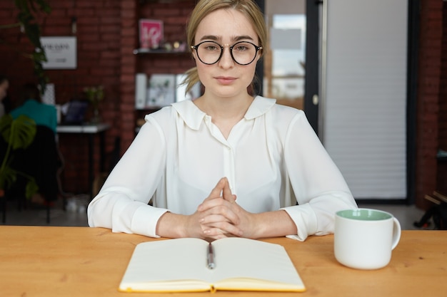 Concepto de personas, negocios, estilo de vida y ocupación. Mujer joven seria especialista en recursos humanos con anteojos redondos y blusa blanca estrechando las manos durante la entrevista de trabajo, sentado en el escritorio y tomando notas