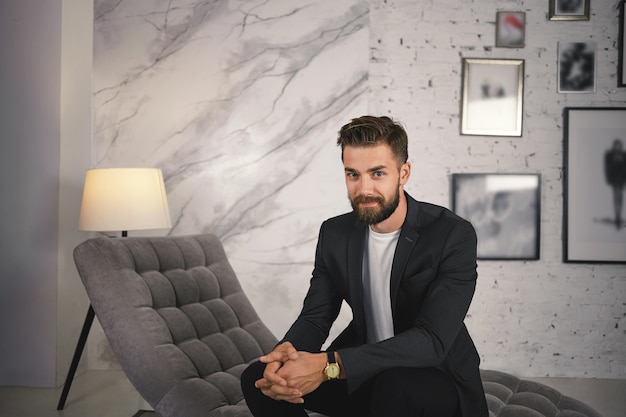 Concepto de personas, negocios, éxito, moda y estilo. Retrato de moda joven empresario europeo exitoso con barba difusa sentado en la moderna sala de estar, vistiendo traje y reloj de pulsera