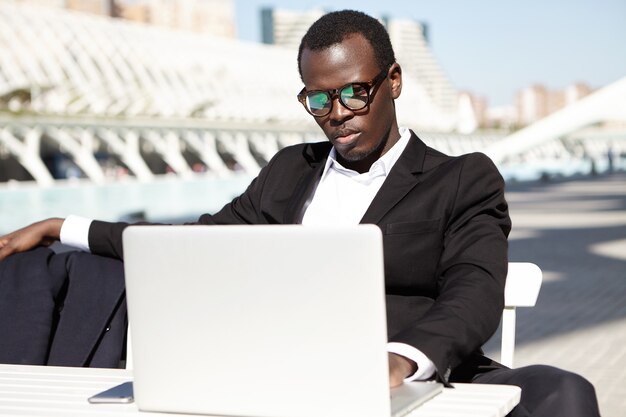 Concepto de personas, negocios, ocupación y tecnología. Hombre serio concentrado en gafas vestido formalmente escribiendo algo en la computadora portátil o leyendo noticias en línea mientras está sentado en la cafetería al aire libre