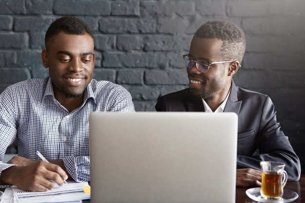 Concepto de personas, negocios, trabajo en equipo y cooperación. Dos trabajadores corporativos afroamericanos en ropa formal trabajando juntos en una presentación común en una computadora portátil genérica en la oficina moderna