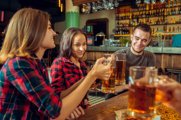 Concepto de personas, ocio, amistad y comunicación: amigos felices bebiendo cerveza, hablando y tintineando vasos en el bar o pub