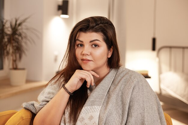 Concepto de personas, ocio, comodidad y estilo de vida. Imagen interior de atractiva mujer joven y bonita de talla grande con cabello negro suelto relajándose en casa, sentada en un sillón, envuelta en una manta cálida