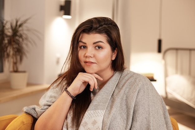 Concepto de personas, ocio, comodidad y estilo de vida. Imagen interior de atractiva mujer joven y bonita de talla grande con cabello negro suelto relajándose en casa, sentada en un sillón, envuelta en una manta cálida