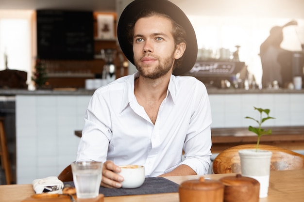 Concepto de personas, ocio y estilo de vida. Filmación en interiores de hombre barbudo joven guapo con sombrero de moda sentado en la mesa de madera con taza, disfrutando de un capuchino fresco durante la pausa para el café, con mirada feliz