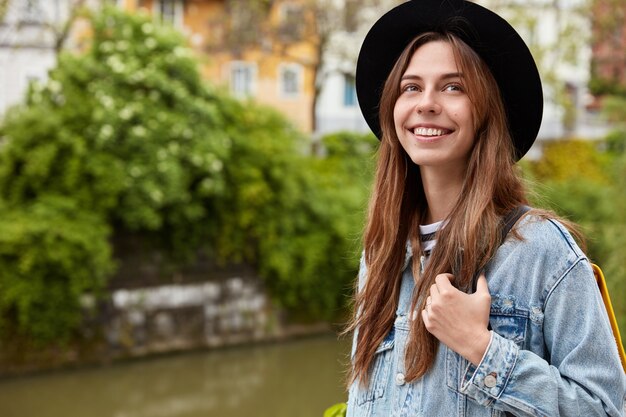 Concepto de personas y ocio. Mujer joven positiva con cabello oscuro, viste sombrero negro de moda, chaqueta vaquera, disfruta de hacer turismo