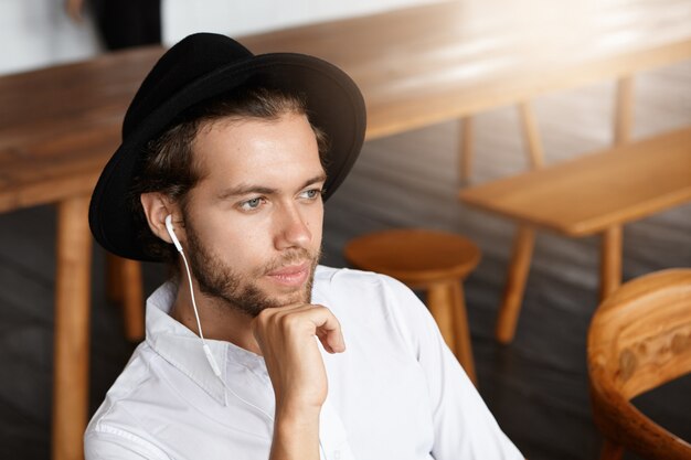 Concepto de personas, ocio y tecnología. Hombre joven de moda con barba relajándose solo en el interior, soñando despierto y escuchando pistas de música en auriculares a través de la aplicación en línea en su dispositivo electrónico