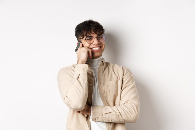 Concepto de personas reales. Chico joven feliz hablando por teléfono móvil, mirar a un lado y sosteniendo el teléfono inteligente cerca de la oreja, de pie sobre fondo blanco.