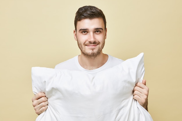 Concepto de personas, ropa de cama y descanso. Retrato de atractivo joven feliz con barba posando aislada sosteniendo almohada blanca yendo a dormir, con sonrisa positiva, diciendo buenas noches