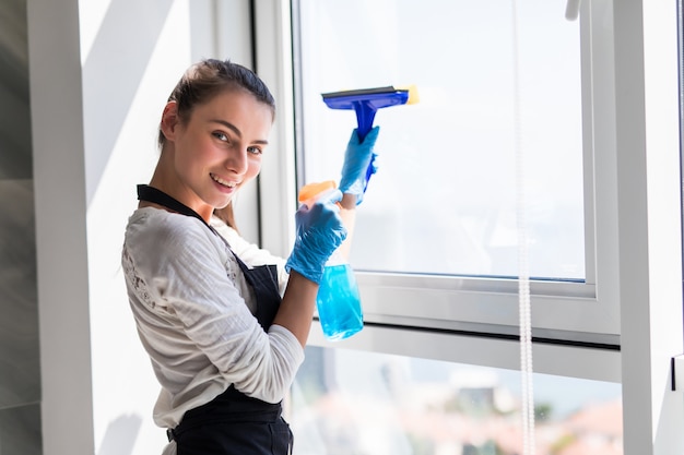 Concepto de personas, tareas domésticas y limpieza. Mujer feliz en guantes de limpieza de ventanas con trapo y spray limpiador en casa