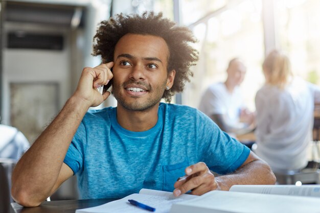 Concepto de personas, tecnología y comunicación. Apuesto estudiante afroamericano con barba sonriendo, teniendo una agradable conversación telefónica