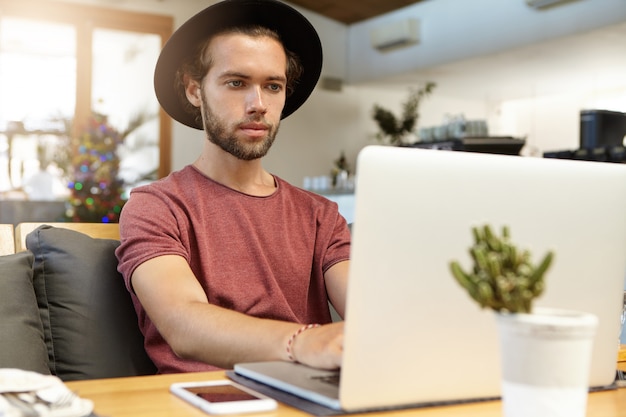 Concepto de personas, tecnología y comunicación. Grave profesional independiente con barba y elegante sombrero sentado frente a una computadora portátil abierta y trabajando de forma remota, utilizando la conexión gratuita a Internet de alta velocidad en el café