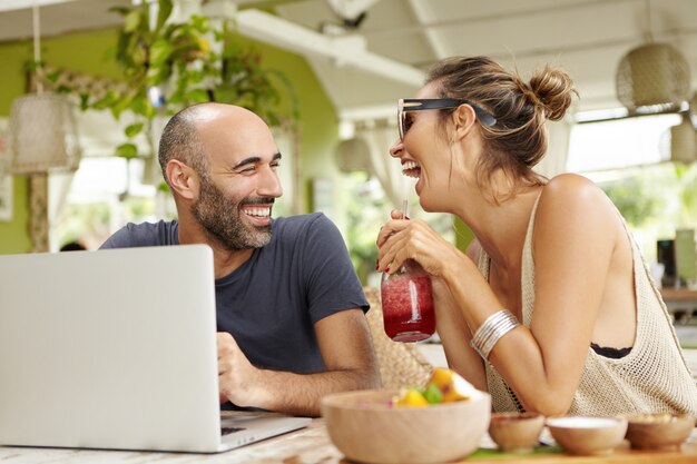 Concepto de personas, tecnología y estilo de vida. Atractivo hombre calvo con barba sentado frente a la computadora portátil y contando chistes a su novia con batido en sus manos.