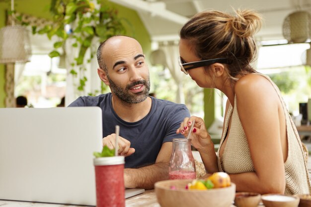 Concepto de personas y tecnología. Hermosa pareja teniendo una agradable conversación, sentado en la mesa de café con portátil y batido en vacaciones de verano.