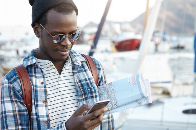 Concepto de personas, tecnología moderna, comunicación, viajes y turismo. Apuesto joven mochilero afroamericano con mapa en papel y teléfono celular, mensajes en línea mientras acaba de llegar a una nueva ciudad