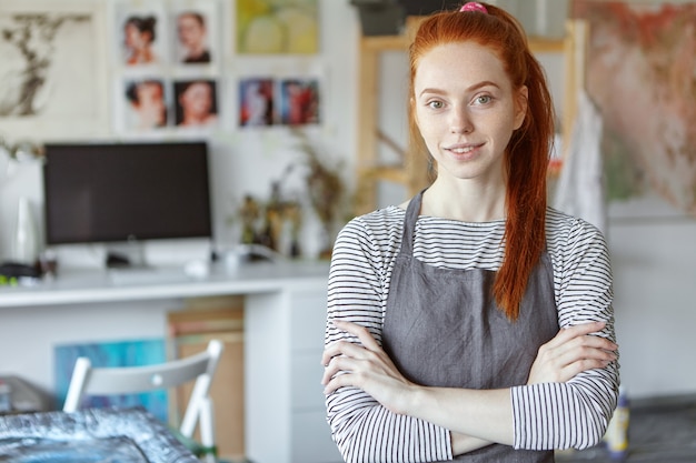 Concepto de personas, trabajo, ocupación, hobby y creatividad. Imagen de mujer joven pelirroja positiva de profesión creativa con delantal sobre top rayado