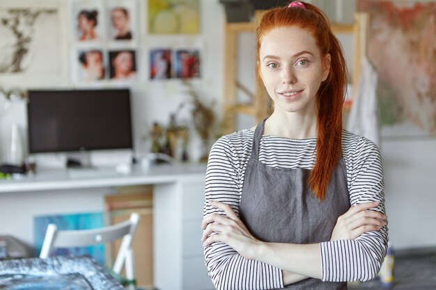 Concepto de personas, trabajo, ocupación, hobby y creatividad. Imagen de mujer joven pelirroja positiva de profesión creativa con delantal sobre top rayado