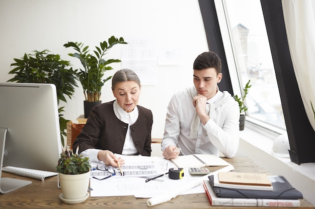 Concepto de personas, trabajo, trabajo en equipo y cooperación. Dos colegas ingenieros profesionales sentados en un escritorio con planos, herramientas informáticas y de ingeniería, discutiendo planes e ideas, con miradas serias