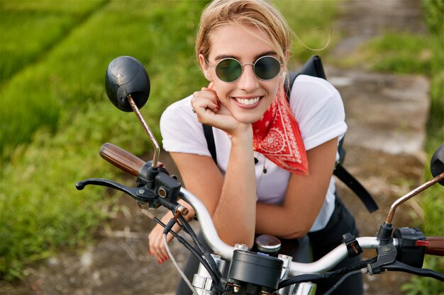 Concepto de personas, transporte y estilo de vida. Feliz joven rubia vestida de manera informal, satisfecha después de un viaje rápido en moto, lleva gafas de sol de moda, sueña con algo agradable