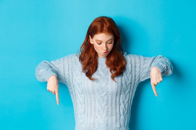 Concepto de personas y vacaciones de invierno. Chica pelirroja intrigada, apuntando y mirando hacia abajo pensativo, haciendo una elección, de pie sobre fondo azul