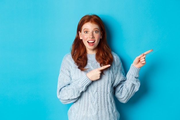 Concepto de personas y vacaciones de invierno. Curiosa adolescente en suéter, señalando con el dedo a la derecha y mirando a la cámara asombrado, mostrando la oferta promocional, de pie sobre fondo azul.