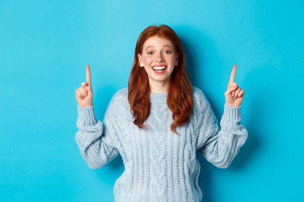 Concepto de personas y vacaciones de invierno. Hermosa adolescente con el pelo rojo, vistiendo un suéter, apuntando con el dedo hacia el logo y sonriendo, mostrando publicidad, fondo azul.