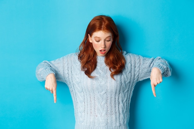 Foto gratuita concepto de personas y vacaciones de invierno. impresionada chica pelirroja en suéter, mirando y apuntando hacia abajo con asombro, de pie contra el fondo azul.