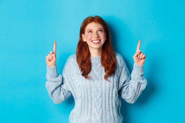 Concepto de personas y vacaciones de invierno. Linda chica adolescente pelirroja apuntando con el dedo hacia arriba, mirando la promoción superior y sonriendo divertido, de pie sobre fondo azul.