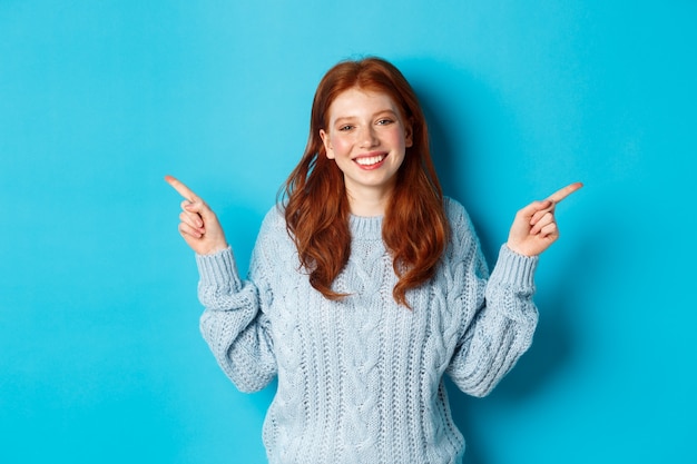 Concepto de personas y vacaciones de invierno. Linda chica adolescente con el pelo rojo, sonriendo y señalando con el dedo hacia los lados, mostrando anuncios, de pie sobre fondo azul.