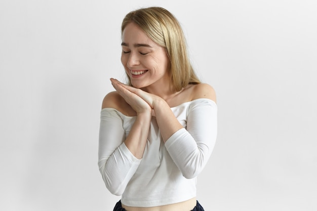 Concepto de positividad, alegría y felicidad. Disparo horizontal de una hermosa joven emocional con cabello rubio suelto sonriendo ampliamente y cerrando los ojos, sintiéndose feliz y agradecida porque ganó en la lotería