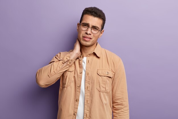 Concepto de problema de salud. Hombre caucásico joven frustrado tiene dolor de cuello, mira con expresión triste, usa camisa marrón, gafas redondas, necesita masaje, inclina la cabeza, aislado sobre una pared púrpura.