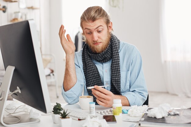 Concepto de problemas de salud. Asombrado joven empleado de oficina con barba tiene un resfriado fuerte, gripe, mira el termómetro con ojos molestos después de medir la temperatura del cuerpo. Gerente enfermo contra el fondo de la oficina