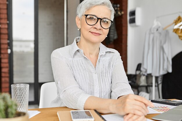 Concepto de profesión, ocupación, trabajo y carrera. Confiado y elegante experto en marketing femenino moderno de unos sesenta años trabajando en la oficina usando computadora portátil, teléfono celular y calculadora, usando anteojos