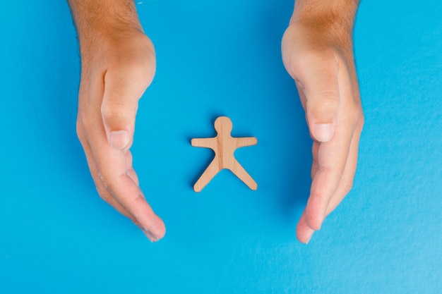 Concepto de protección social en la mesa azul plana lay. Manos cuidando la figura humana de madera.