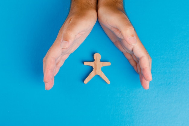 Concepto de protección social en la mesa azul plana lay. Manos cuidando la figura humana de madera.