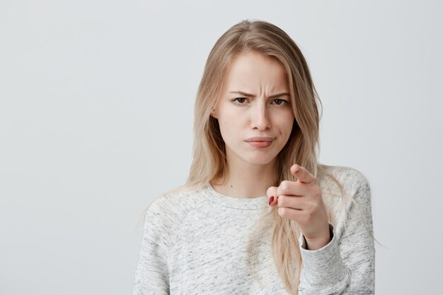 Concepto de publicidad. Emocionada alegre mujer rubia europea con camiseta de manga larga, cejas fruncidas y señalar con el dedo índice