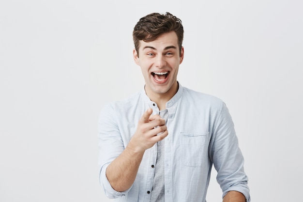 Concepto de publicidad. Emocionado hombre europeo alegre con camisa azul claro que levanta las cejas y exclama, señala con el dedo índice, lo elige, motiva y atrae a los clientes.