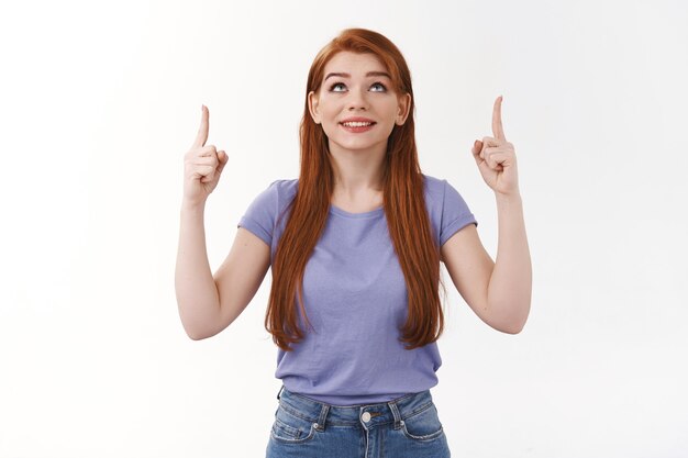 Concepto de publicidad, empleo y emociones. Estudiante pelirroja curiosa soñadora con el pelo largo de jengibre, contemplar algo maravilloso, mirar y señalar divertido, pared blanca