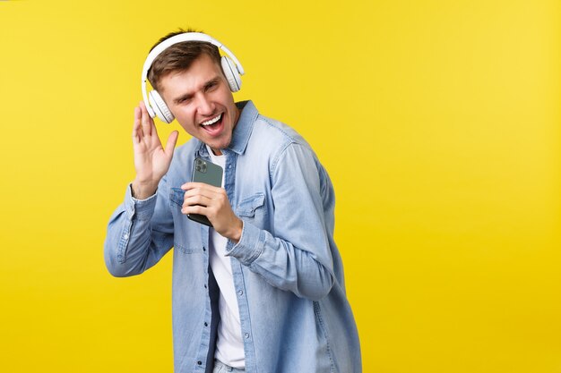 Concepto de publicidad de ocio, tecnología y aplicaciones. Hombre caucásico joven hermoso que se divierte, que juega la aplicación de karaoke en el teléfono móvil, que usa el teléfono inteligente como micrófono y que canta con los auriculares puestos.