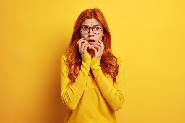 Concepto de reacciones humanas. Mujer preocupada ansiosa mira indignada por tener miedo de algo abre boca agarra cara lleva gafas transparentes y cuello alto.