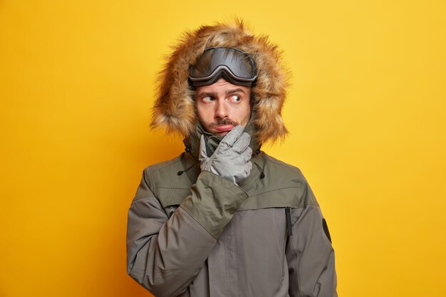 Concepto de recreación y deporte de invierno. Hombre serio pensativo en ropa de abrigo vestido con chaqueta y guantes lleva gafas de snowboard piensa en planes sobre el tiempo libre.